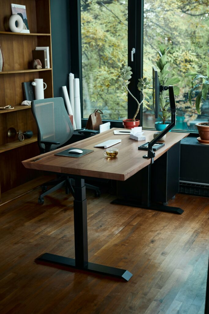 Height-adjustable ergonomics desk with adjustable ergonomic chair, adjustable monitor arm, keyboard, and mouse positioned at a 90 degree angle to a window with lots of green, leafy trees outside