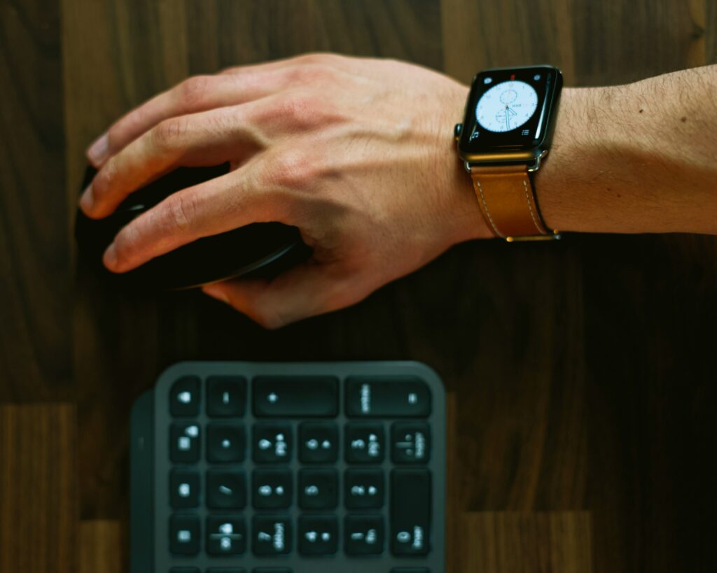 Hand placed on top of computer mouse on right side of keyboard. A smart watch is on the forearm showing displaying an analog clock