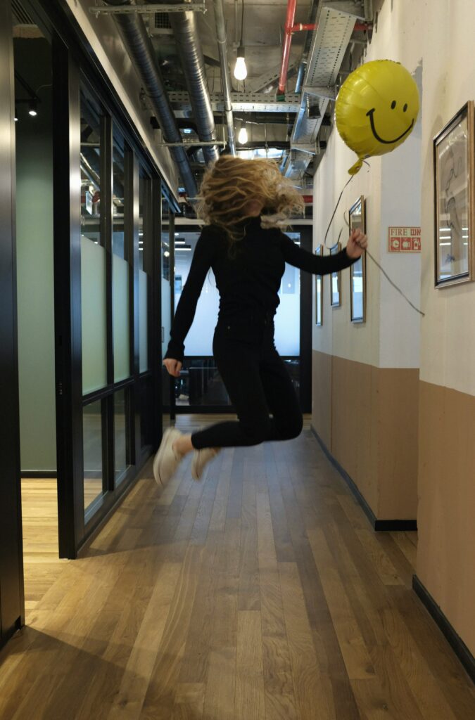 Side view of a woman jumping high in a corridor holding a yellow ballon with smiley face