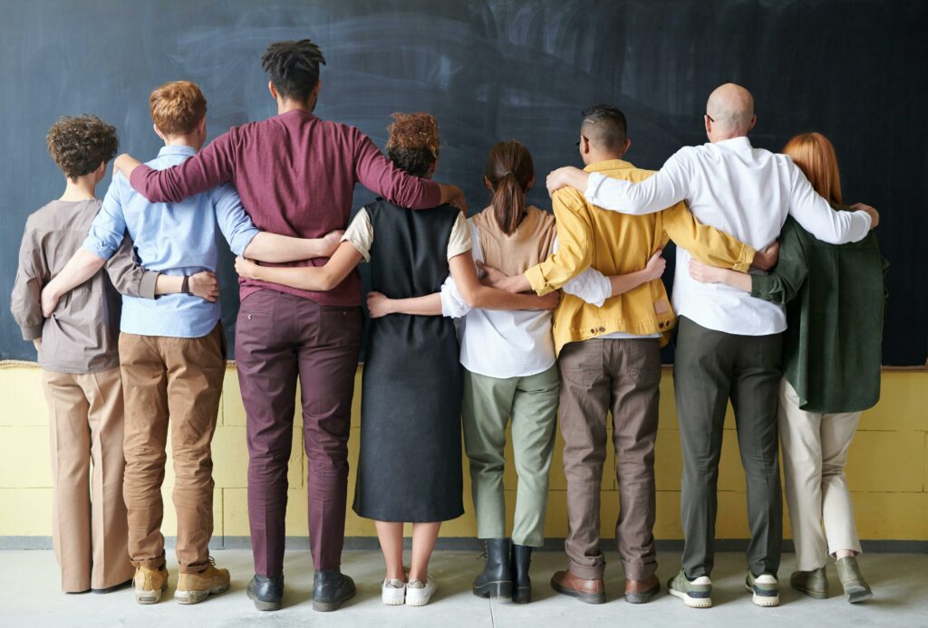 Rear view of eight people of diverse sizes, ages, and genders standing arm-in-arm facing a chalkboard