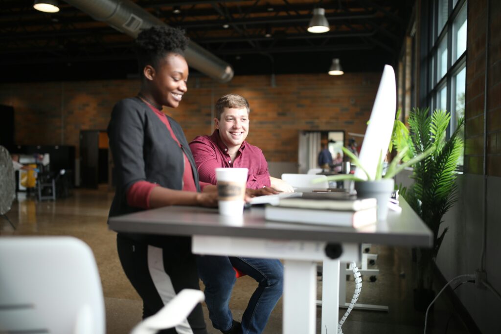 Woman and man standing at electric height-adjustable desk in an open office area. They are looking at a monitor and smiling.